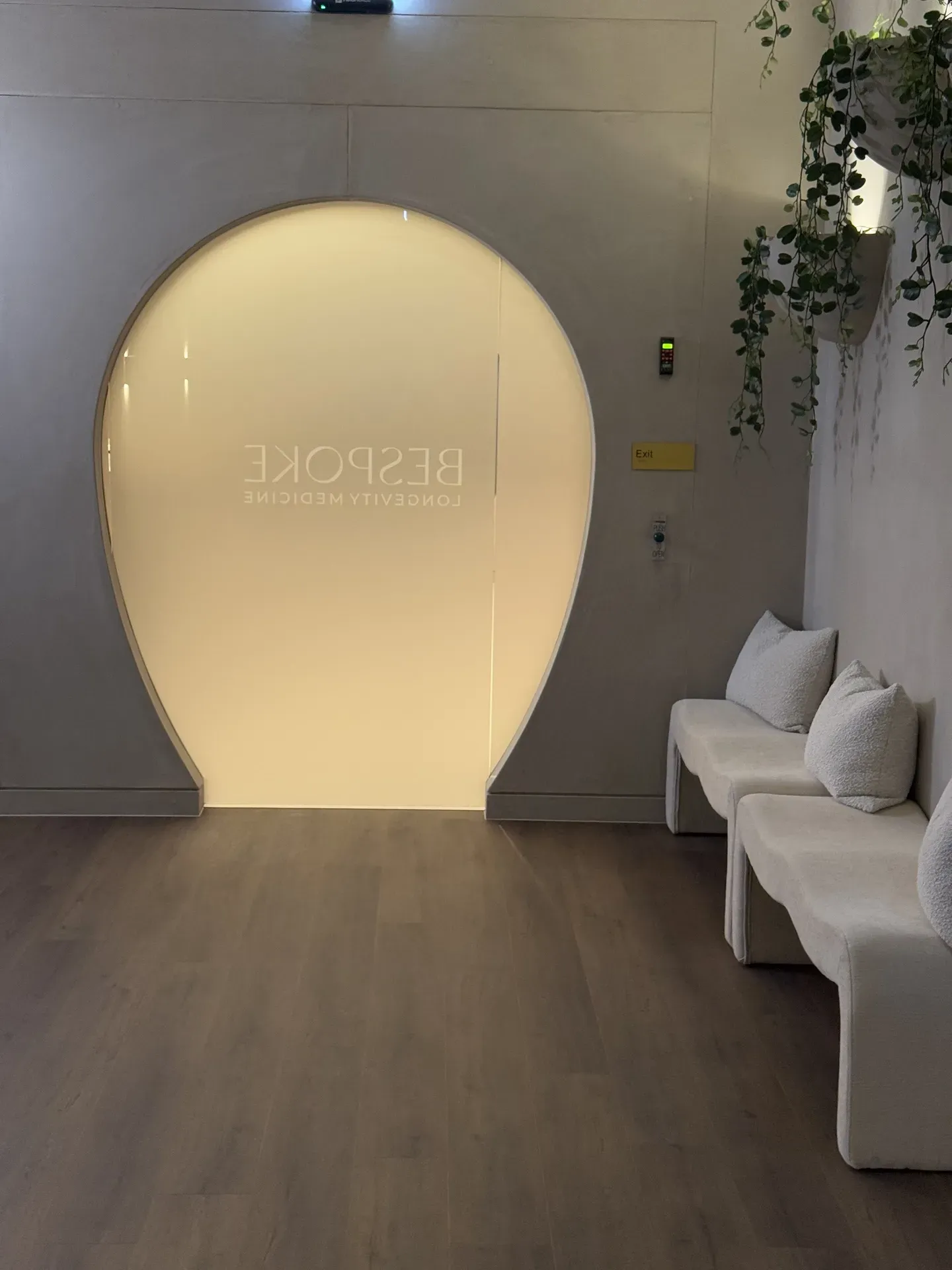Waiting area with curved doorway, benches, and trailing plants. Light walls and wooden floor.