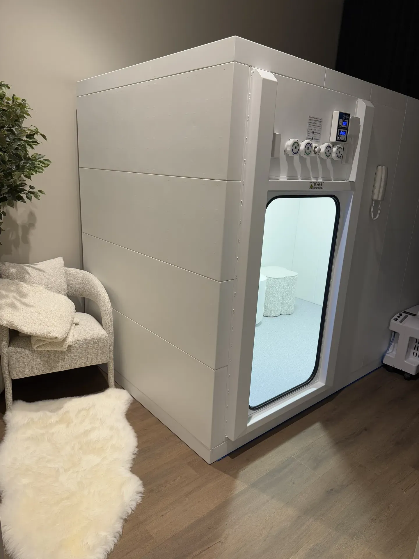 White hyperbaric chamber with porthole door; interior visible. Room with chair and rug.