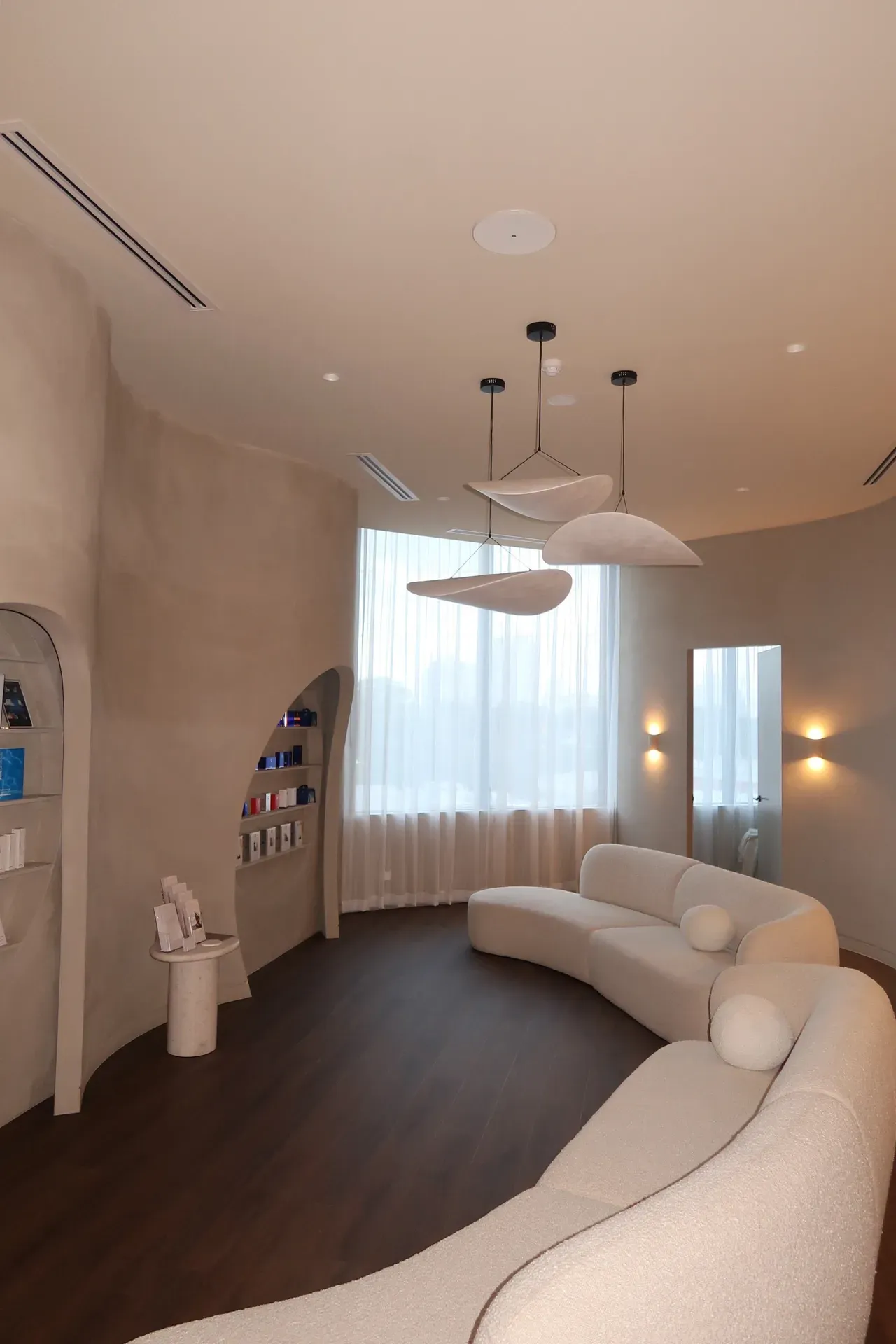 Neutral-toned spa waiting area with curved sofa, large window, and cloud-shaped pendant lights.