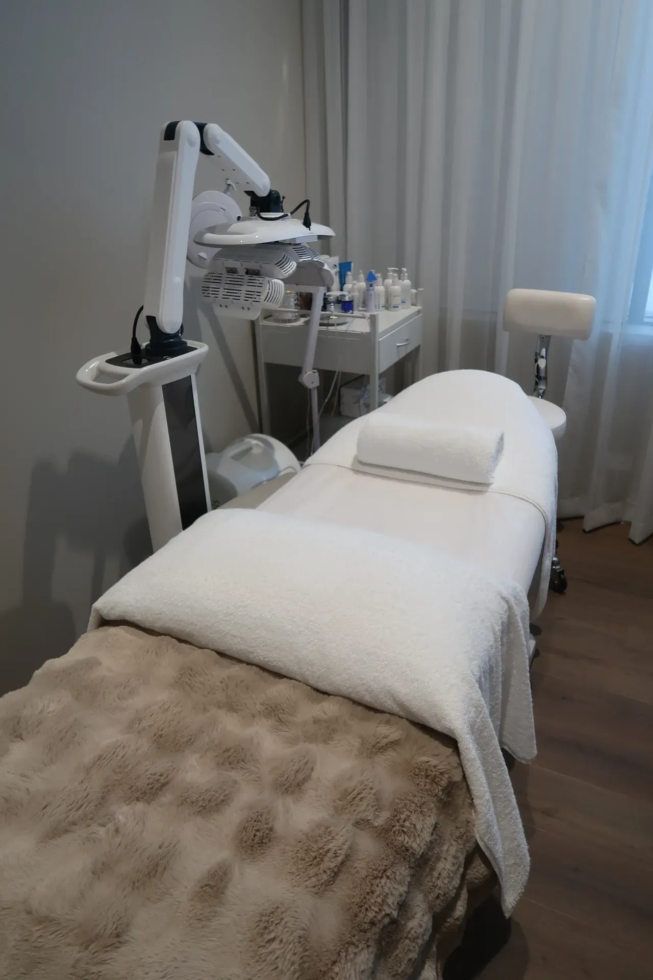 Aesthetician's treatment room: white bed with towel, skincare equipment, small table with products, and a stool.