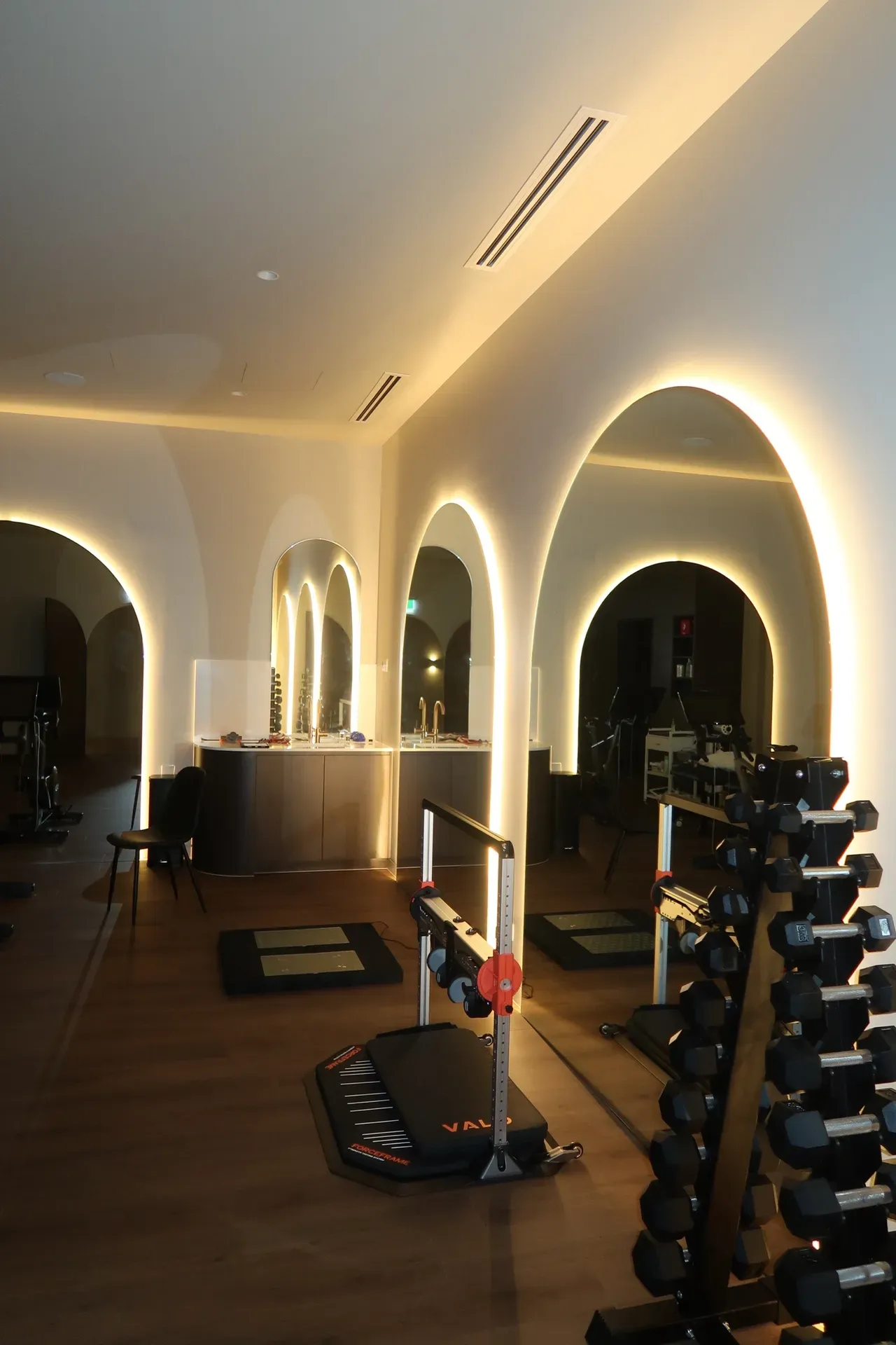 Gym interior with arched mirrors, weights, and workout equipment. Warm lighting. Brown floor.