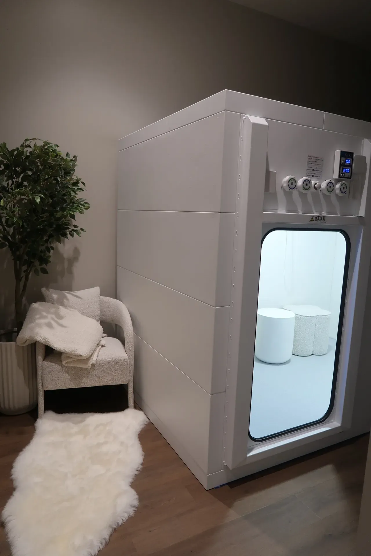 White floatation therapy pod, chair, rug, and plant in a room. The pod is open.