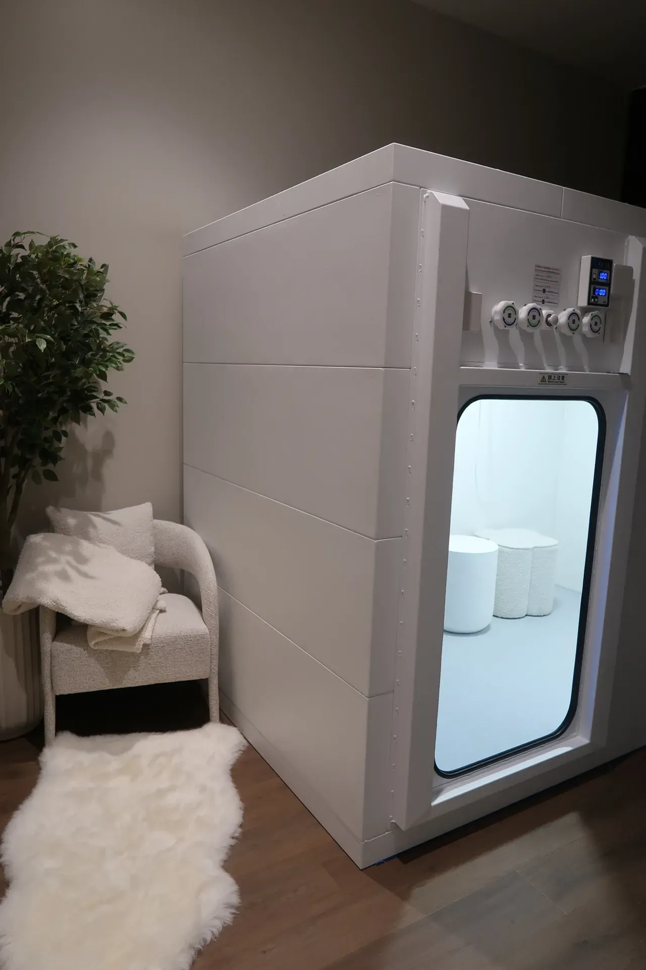 White rectangular floatation therapy pod with an open door in a room with a chair, rug, and plant.