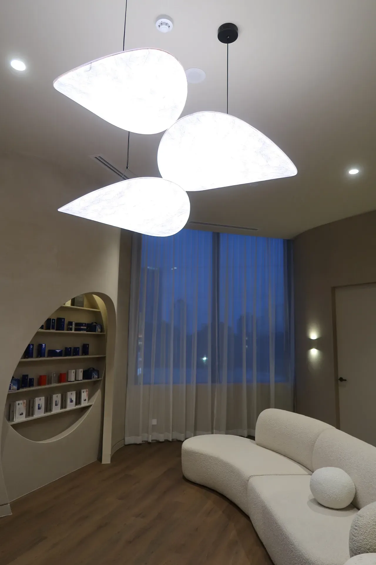 Modern waiting room with curved white sofa, teardrop-shaped lights, and window with sheer curtains.