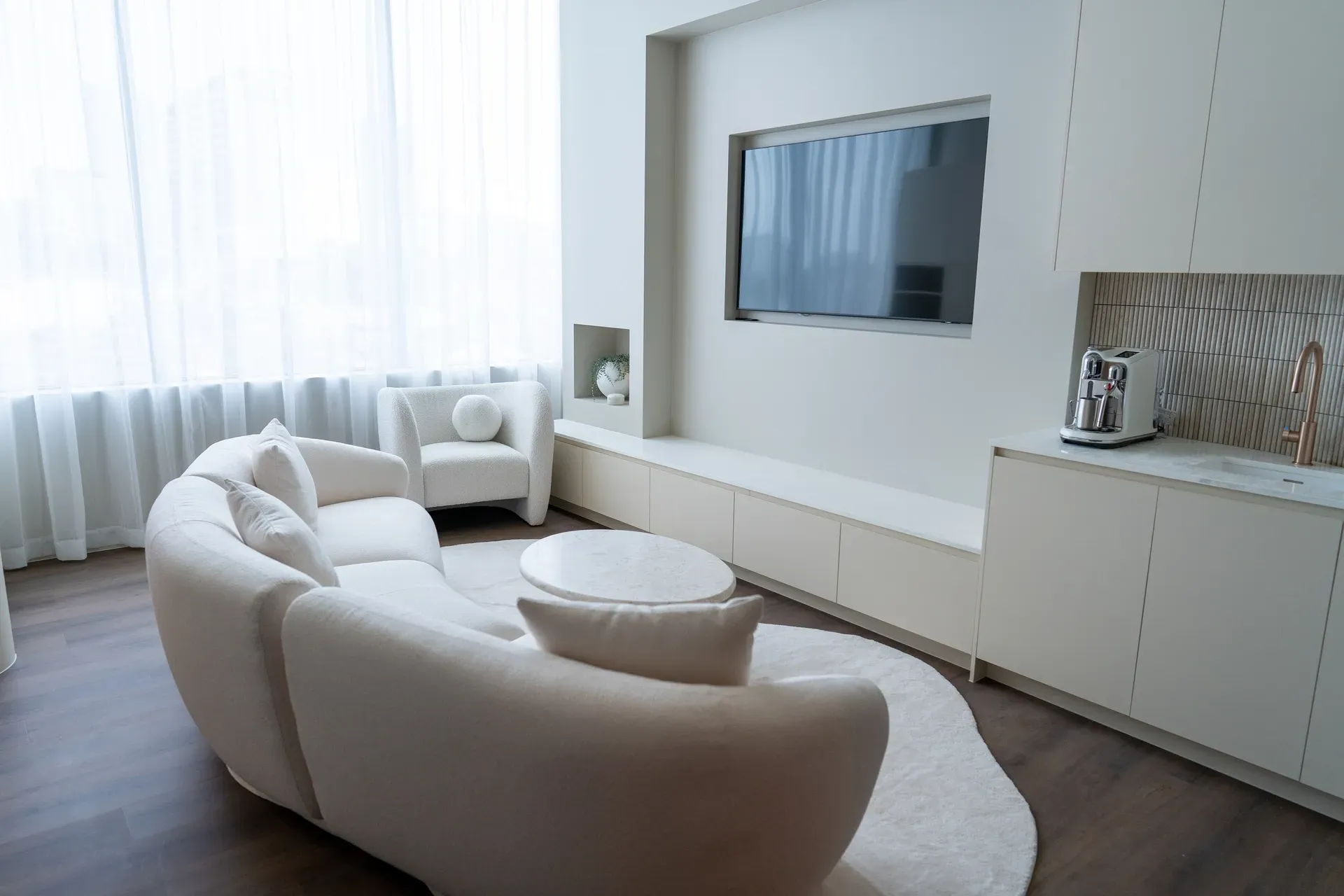 Cream-colored modern living room with curved sofa, round rug, TV, and built-in storage.