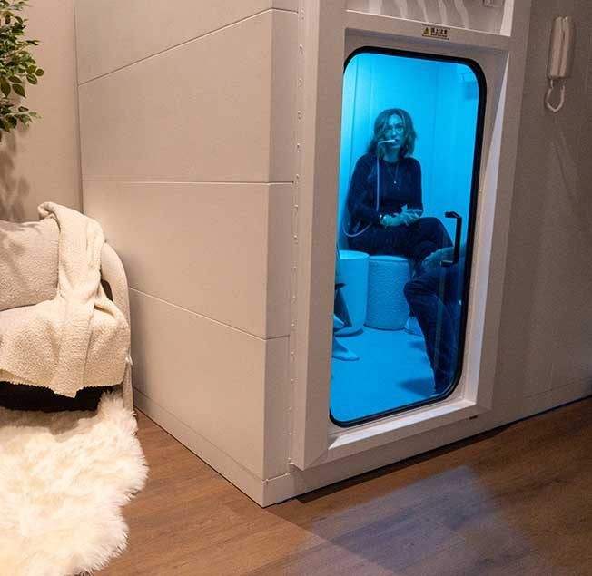 Woman inside a white cold therapy pod with blue lighting. Adjacent is a living room chair and rug.