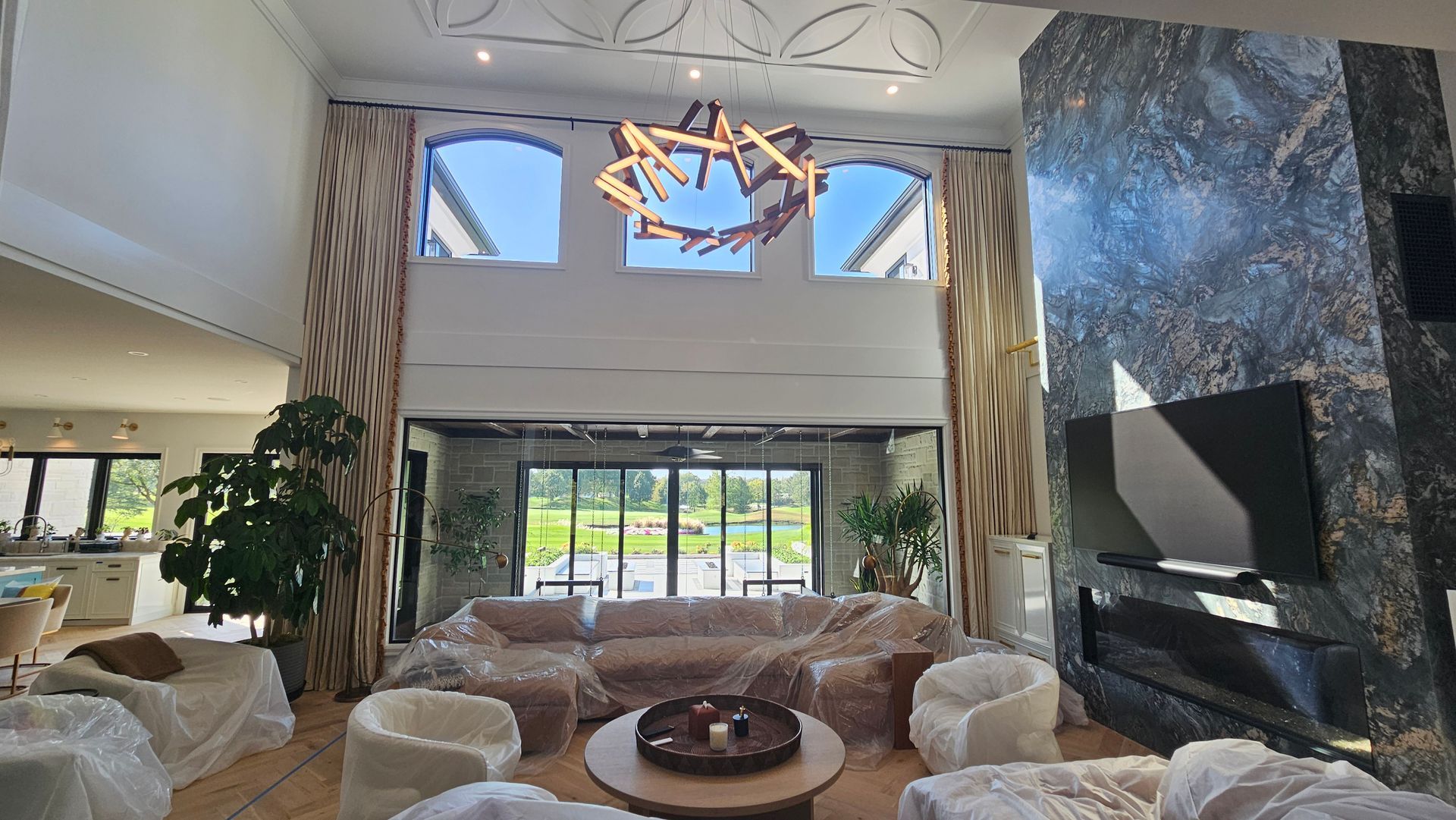 Spacious living room with large windows, a modern fireplace, and furniture covered in plastic.