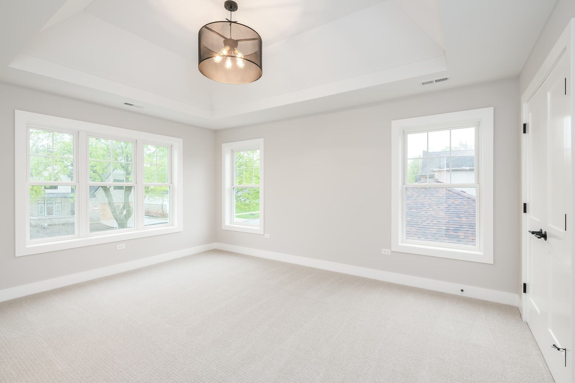 Empty bedroom with white walls, carpet, and windows. A dark light fixture hangs from the ceiling.