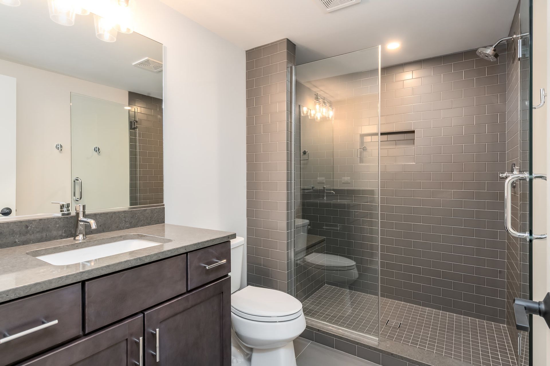 Modern bathroom with gray tiles, glass shower, dark vanity, and white toilet.