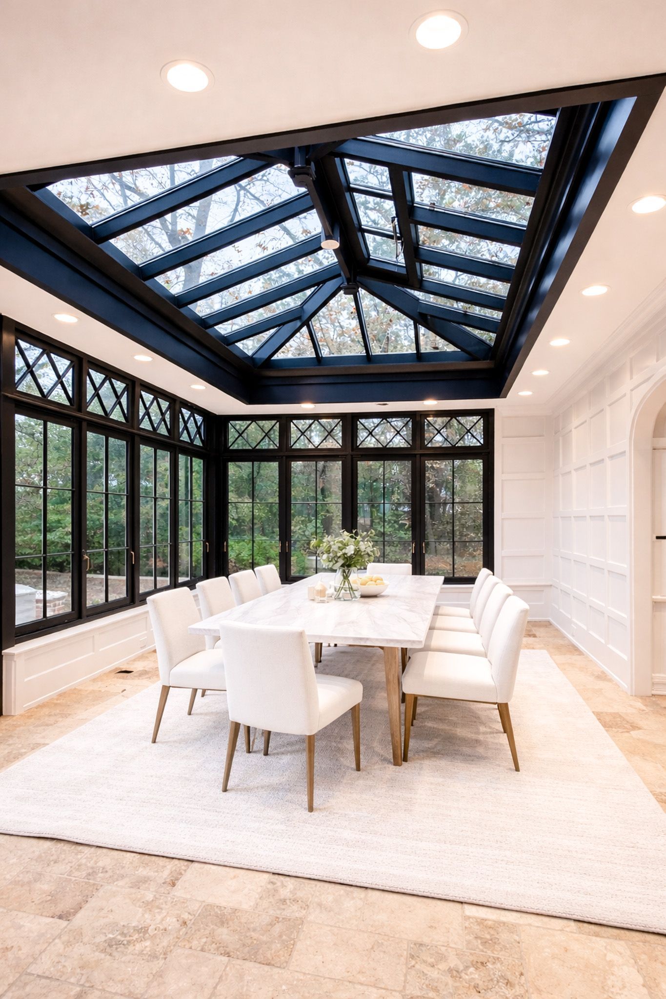 Dining room with wooden table and chairs, a tufted bench, and a crystal chandelier by large windows.