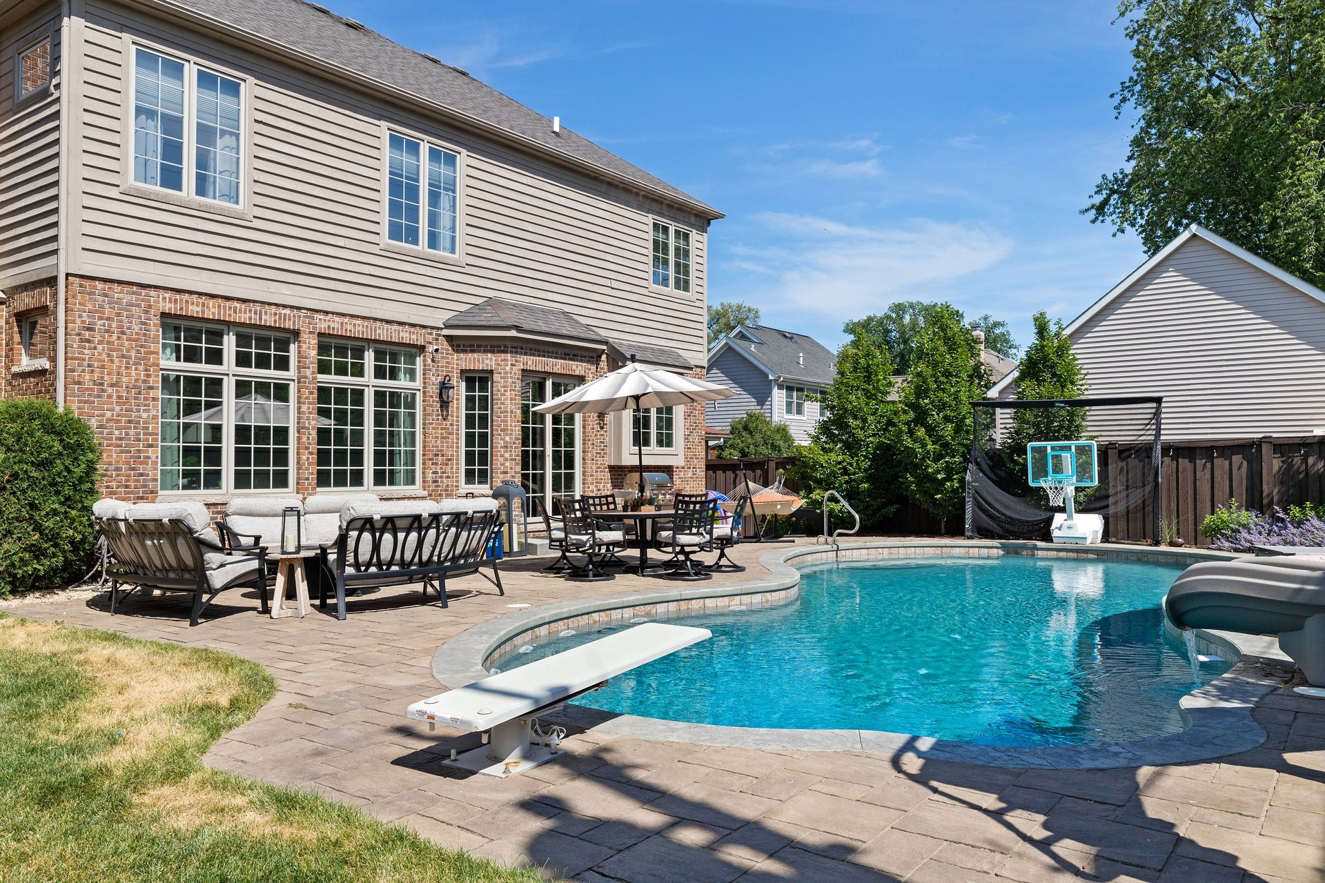 Two-story house with a pool and patio. Brick and siding exterior. Pool is blue, sunny day. Outdoor furniture.