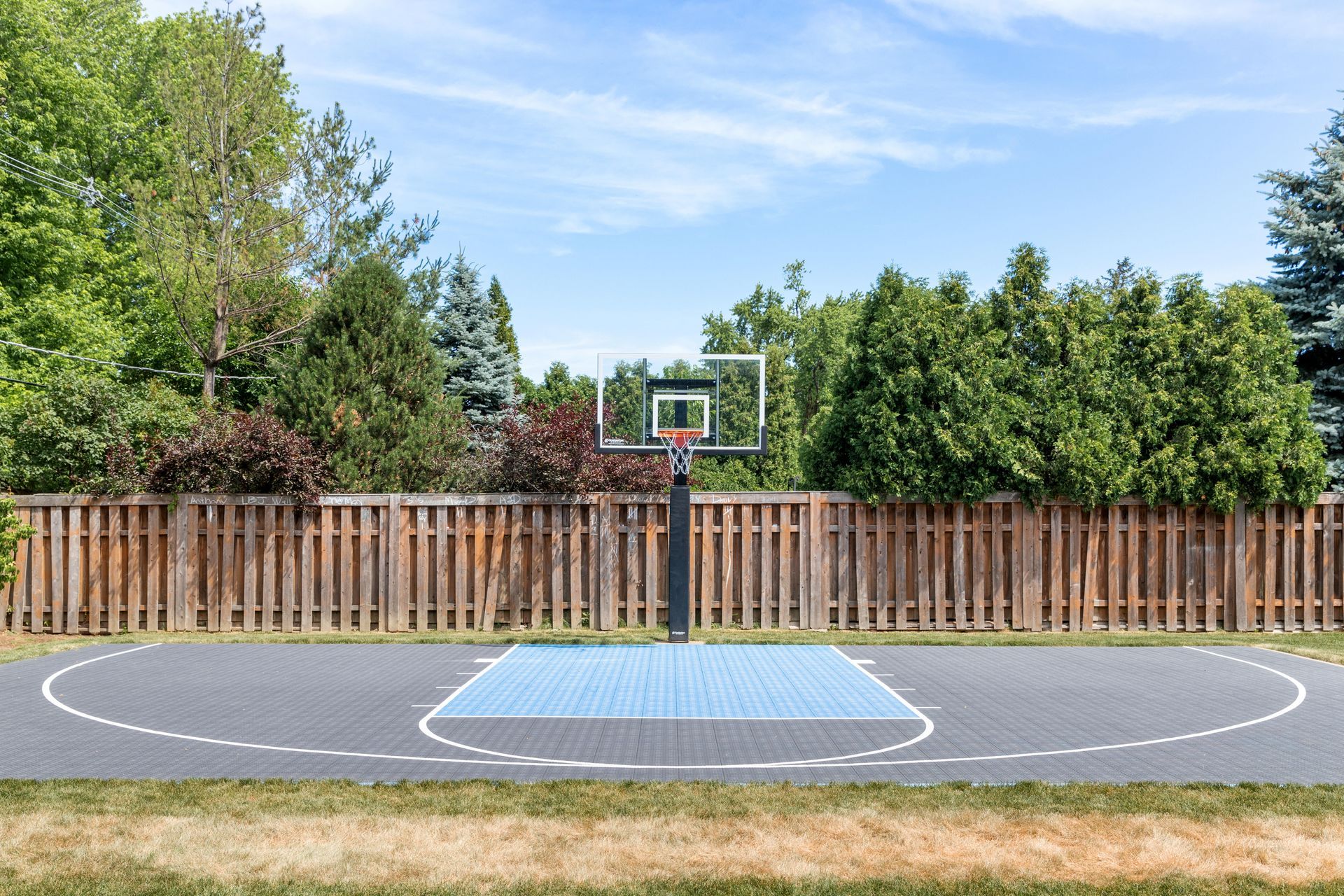 Basketball court in a backyard with a hoop, fence, and trees against a blue sky.