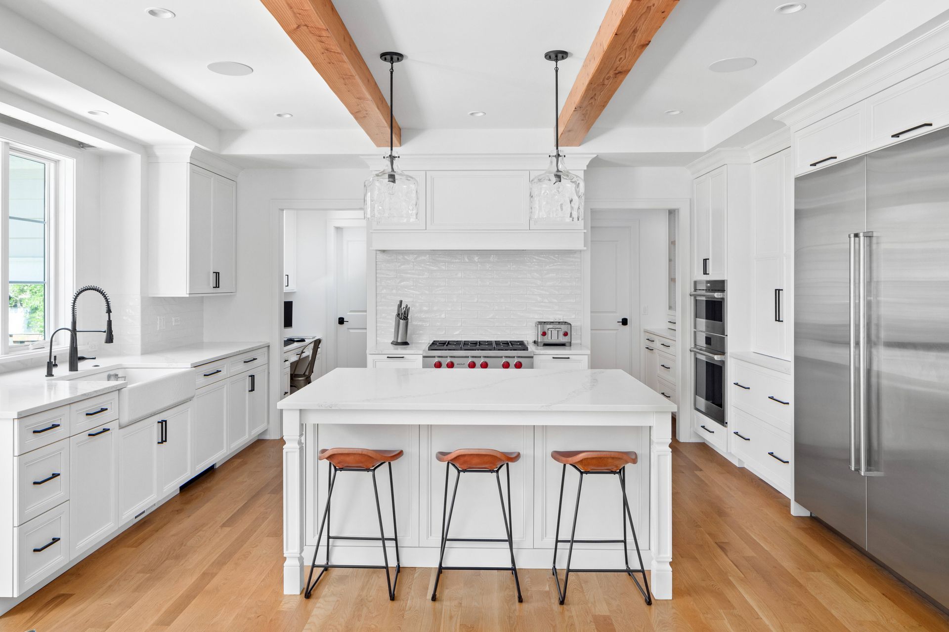 White kitchen with wooden beams, island with stools, stainless steel appliances, and hardwood floors.