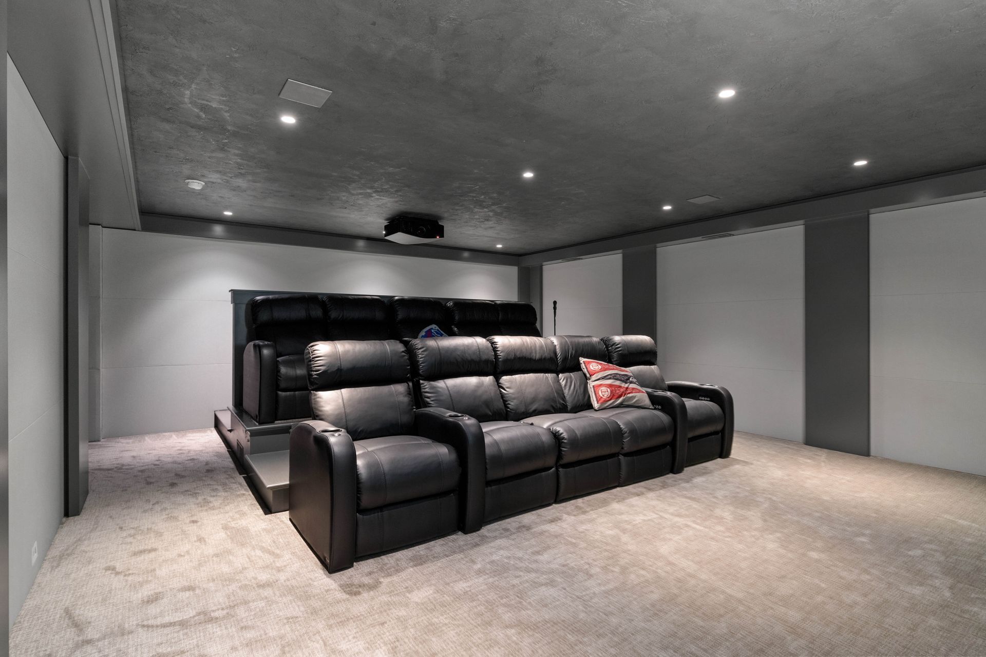 Home theater with black leather recliners, gray walls, and carpet. A projector hangs from the ceiling.