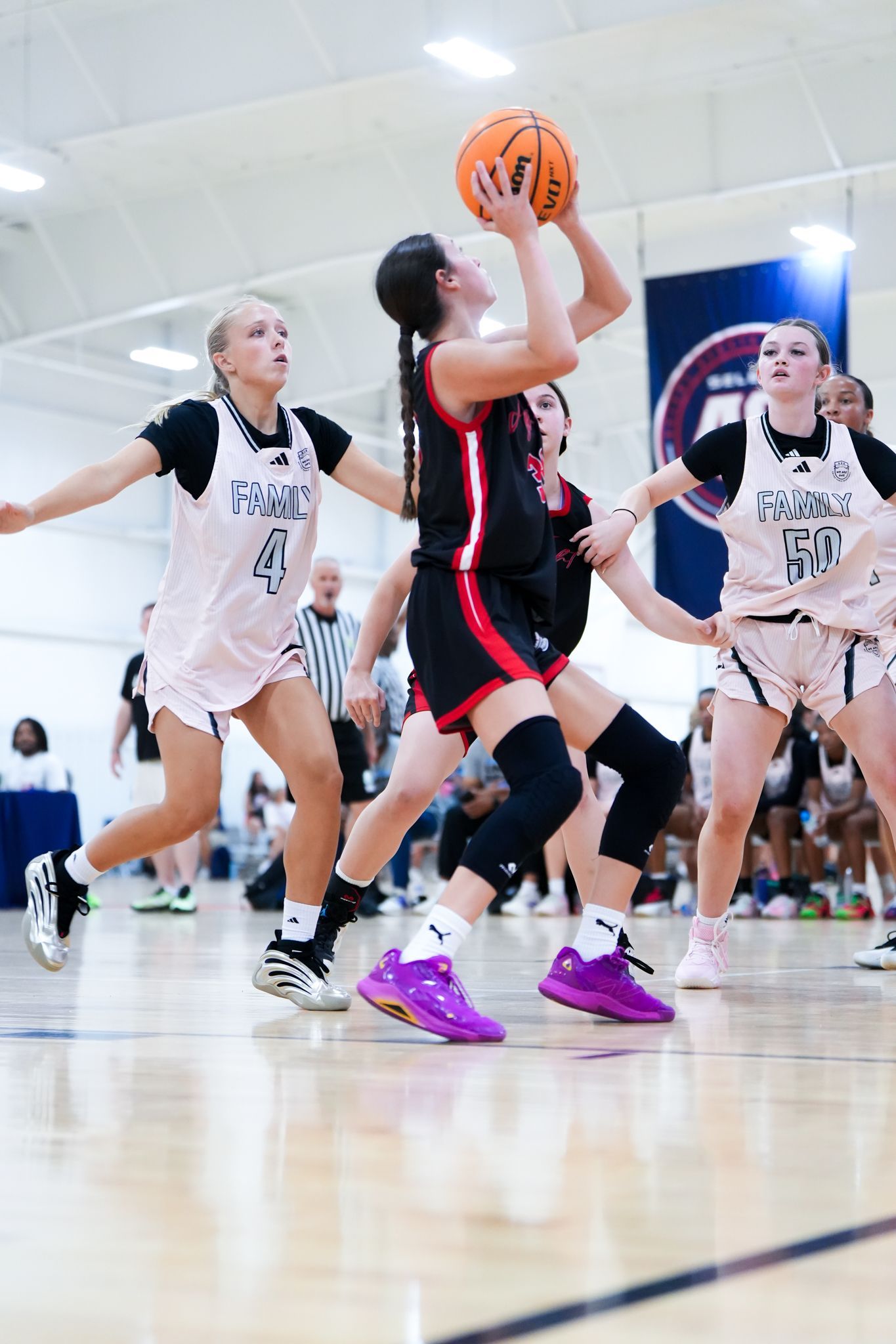 Basketball player in black and red shooting, defended by two in white; indoor court.