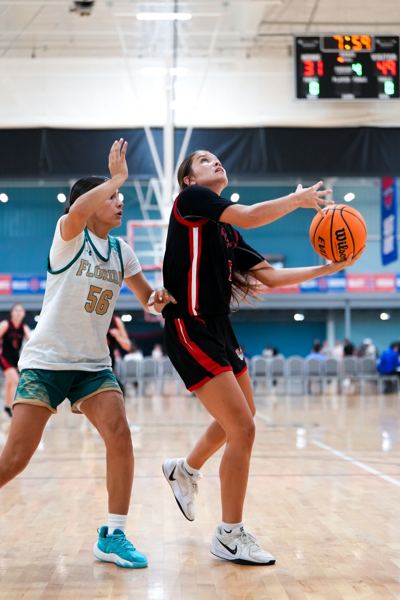 Basketball player in black and red uniform shoots, defended by another player in green. Indoor court.