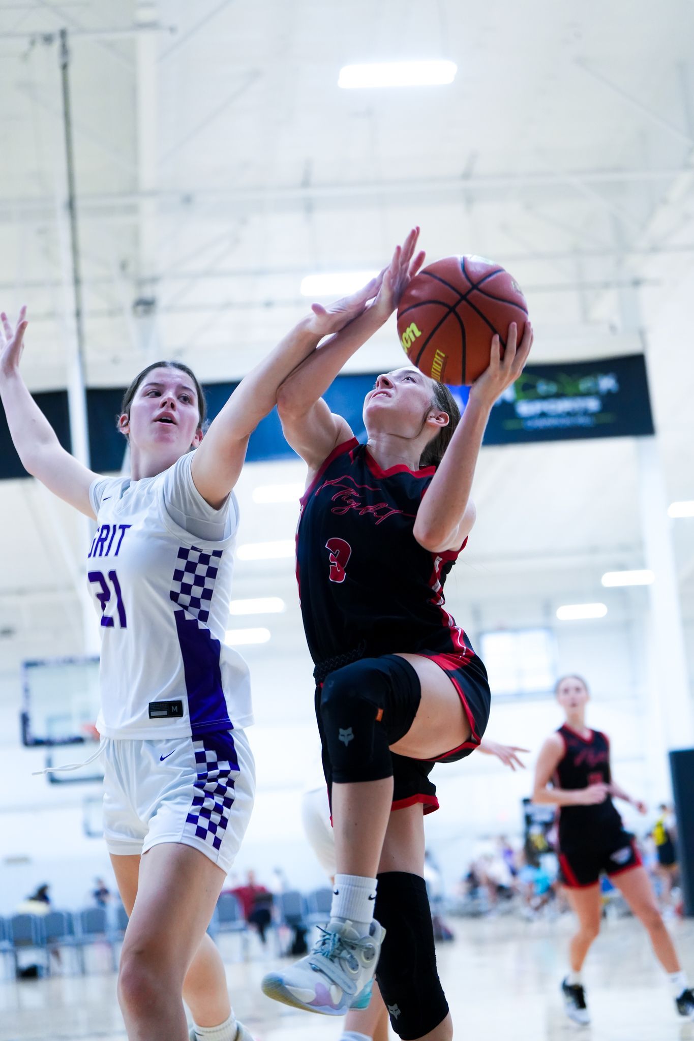 Basketball players in action: one shoots as opponent tries to block, inside a gym.
