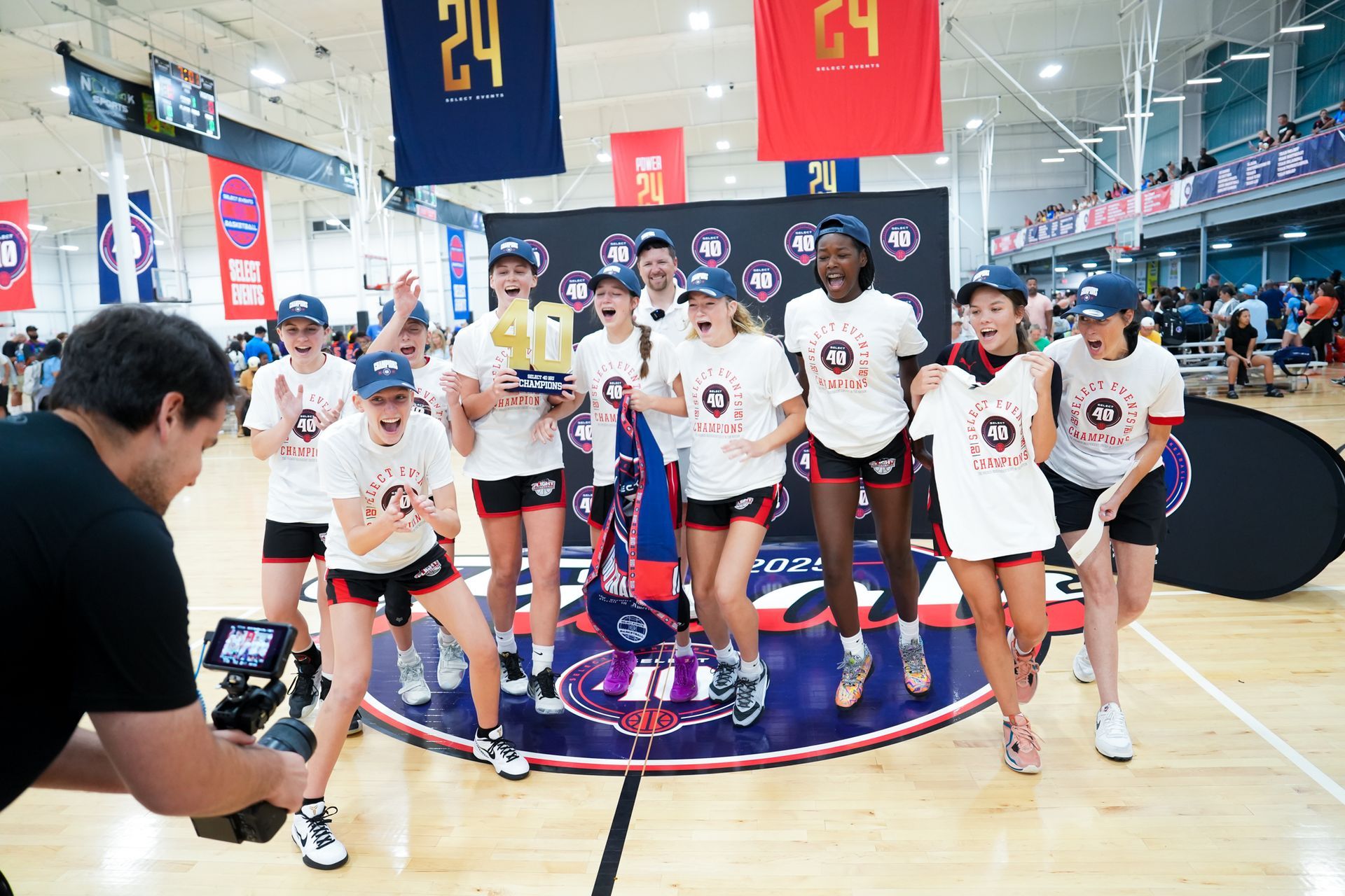 Girls' basketball team celebrating championship win, holding trophy, smiling, indoors, blue and red banners.