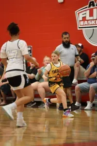 A young girl in yellow and black uniform dribbles a basketball during a game. Referee watches nearby.