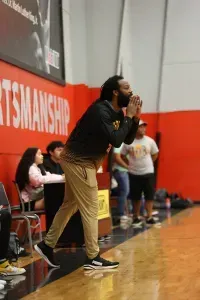 Basketball coach yelling on court; wearing black and tan; spectators in the background.
