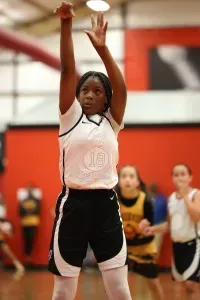 Girl in basketball uniform shooting a free throw, indoors, red background, other players blurred.