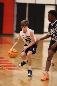 Boy dribbles a basketball during a game; another player defends. Court setting; dark blue and orange colors.