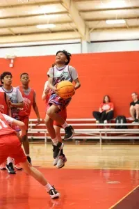 Basketball player jumping to shoot in a red-walled gym. Other players watch; a few spectators sit on bleachers.