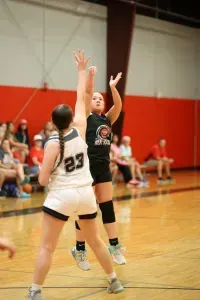 Basketball players in a gym, one shooting while being guarded. Red wall in the background.