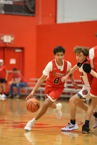 Boy dribbles basketball in a red uniform on a court, other players in the background.