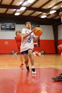 Boy in white jersey with ball dribbles toward the basket; another player in red follows in indoor court.