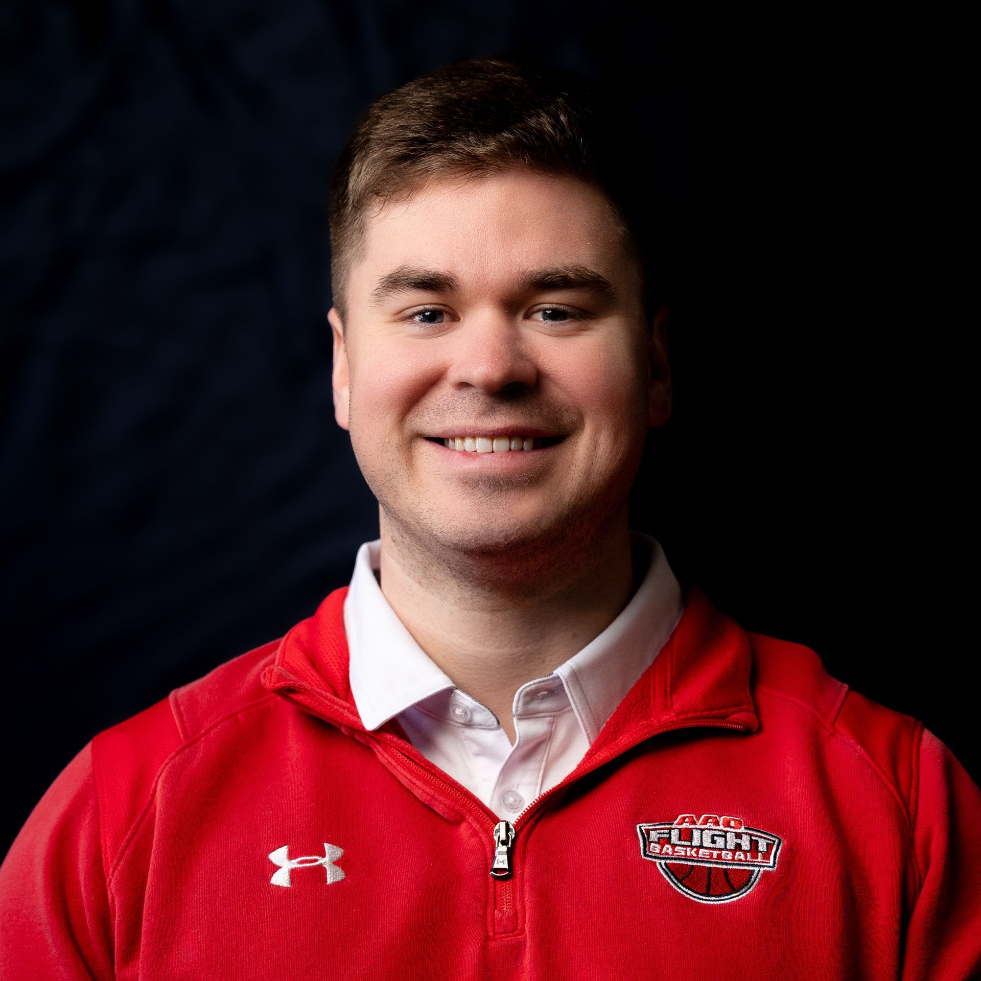 Portrait of a smiling person in a red zip-up jacket against a dark background