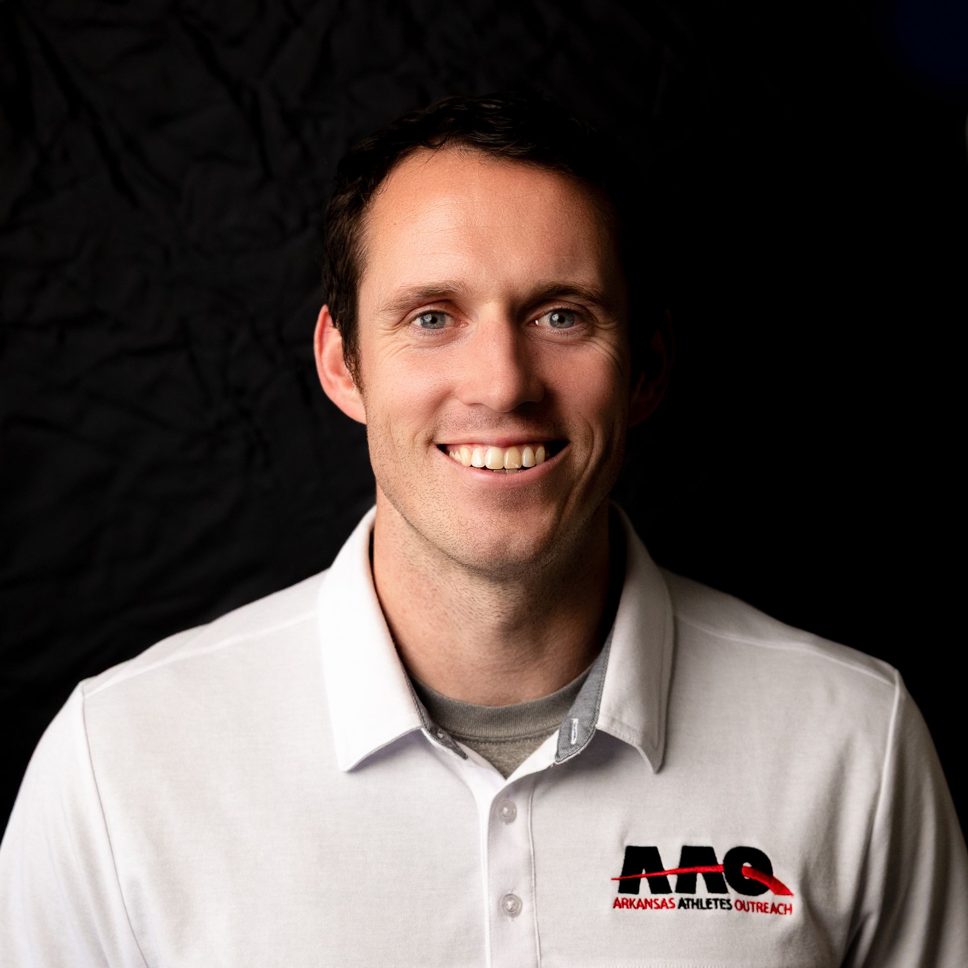 Smiling person in a white polo shirt with a red-and-black logo against a dark background