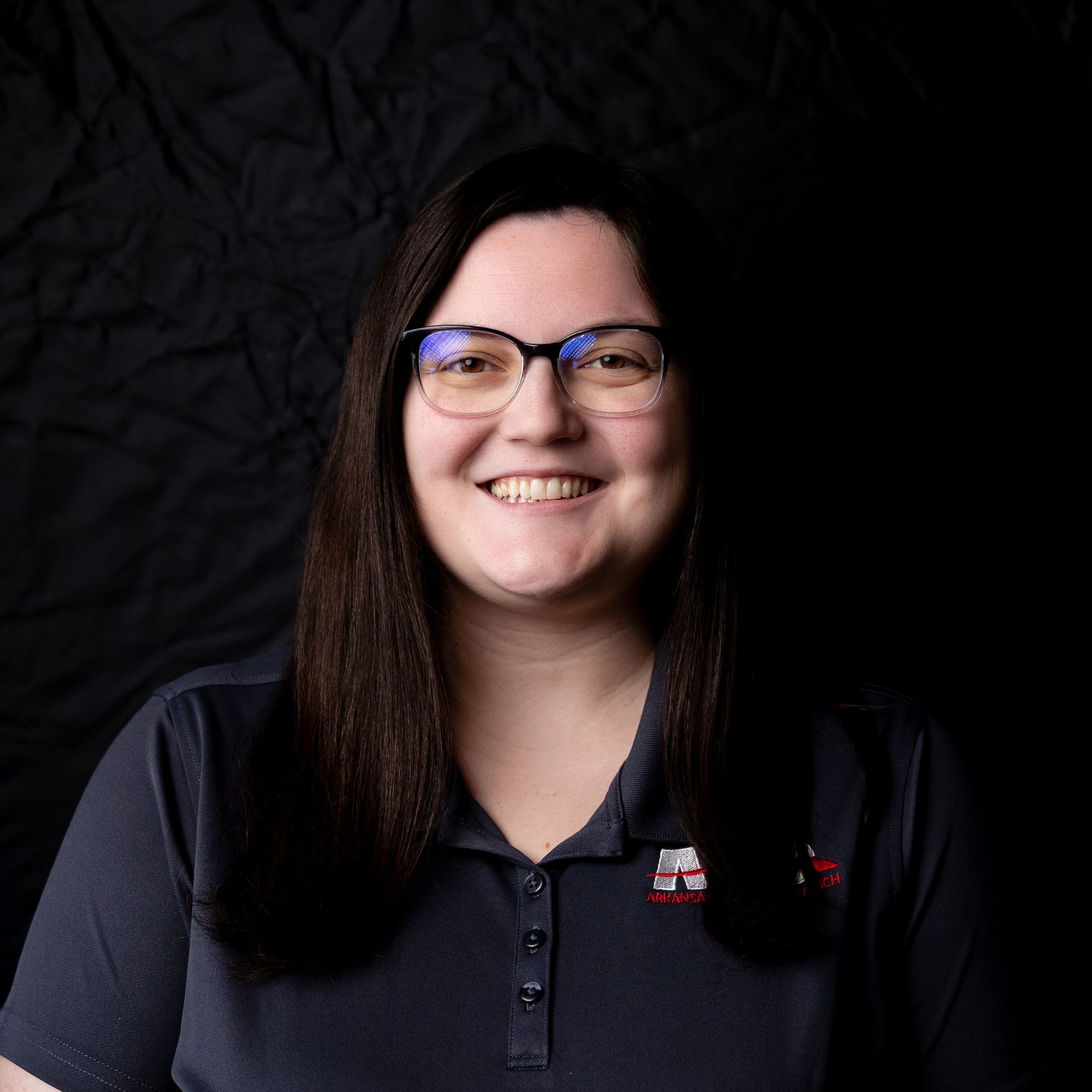 Smiling person with glasses and long dark hair against a black background, wearing a dark polo shirt