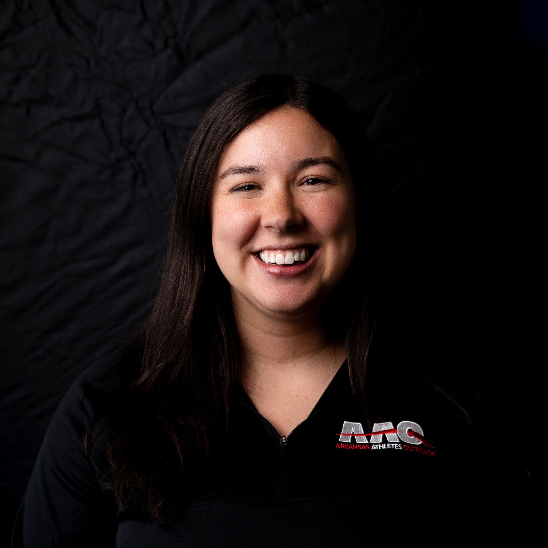 Smiling person in a black shirt with an AEC logo against a dark background