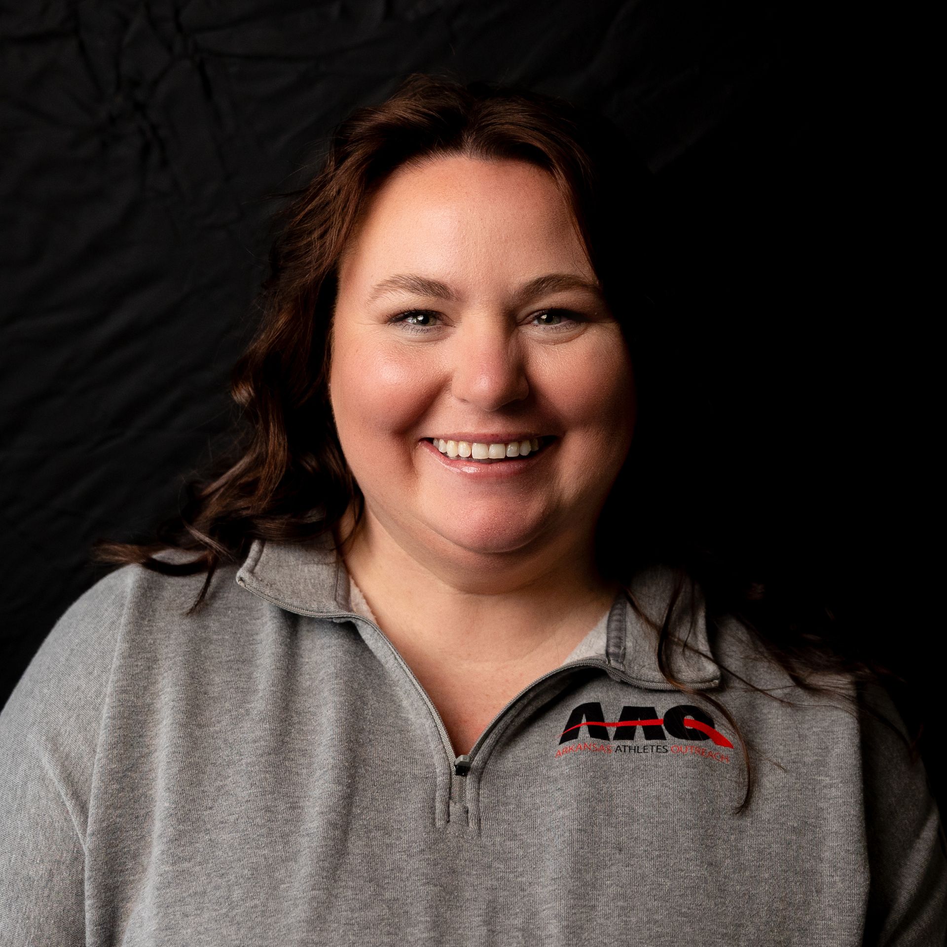 Smiling woman in a gray zip-up shirt with a red logo against a black background