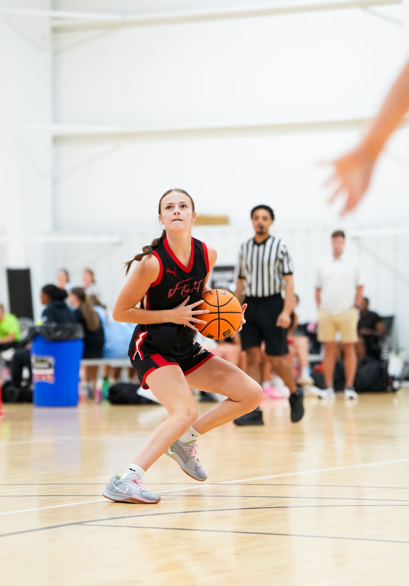 Female basketball player dribbling the ball on court, wearing black and red uniform, referee in the background.