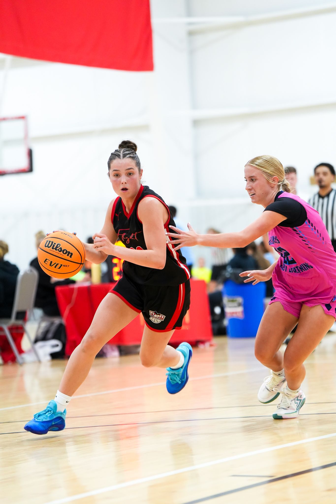Basketball player dribbles the ball past a defender in a game; red and pink uniforms.