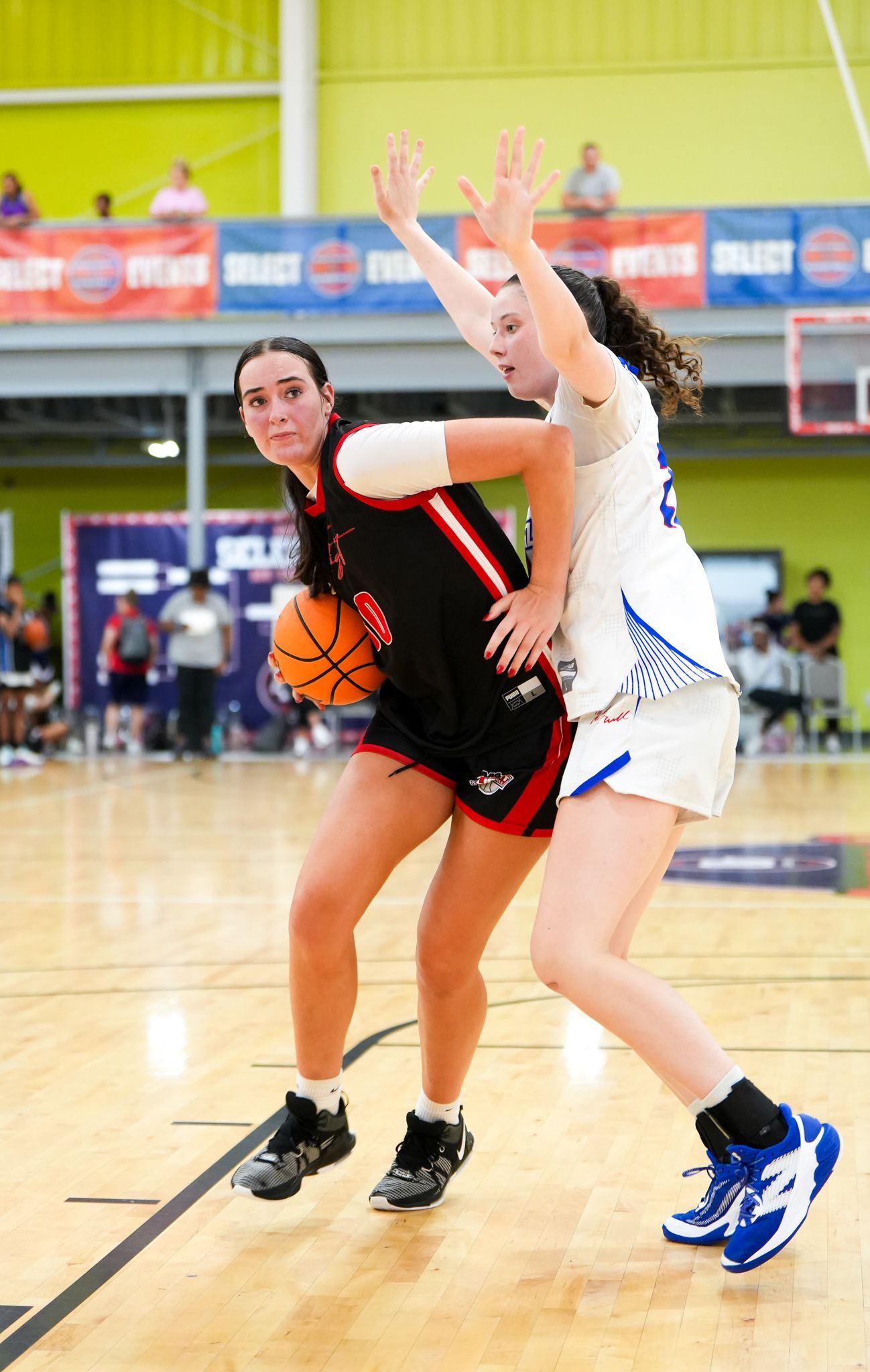 Basketball players, one in black and red dribbles past a defender in blue and white.
