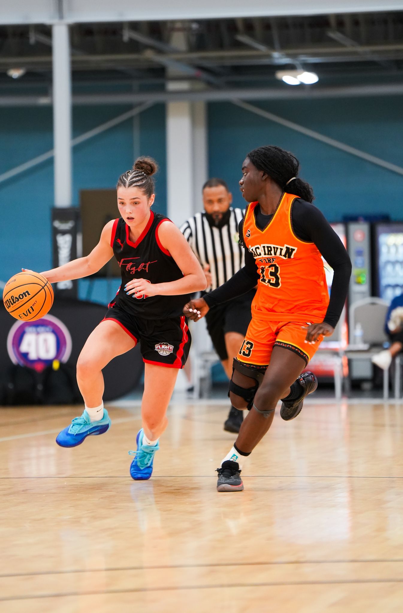Basketball game: two players in action, one dribbling, one defending. Referee nearby.