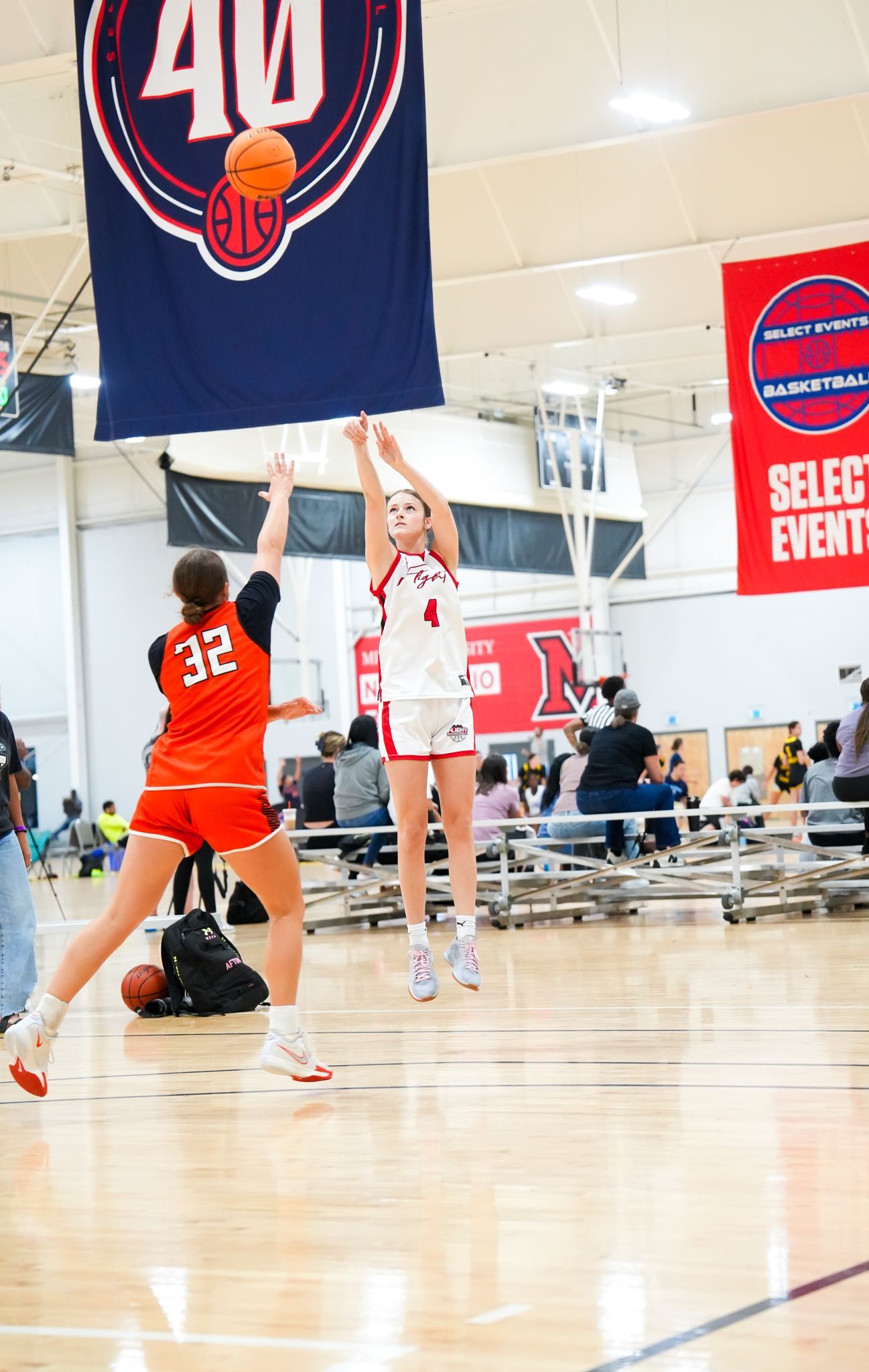 A basketball player shoots, guarded by an opponent. Indoor game, red and orange uniforms. Ball in the air.