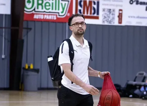 Man in a white polo and glasses with a backpack, holding a basketball in a gym.