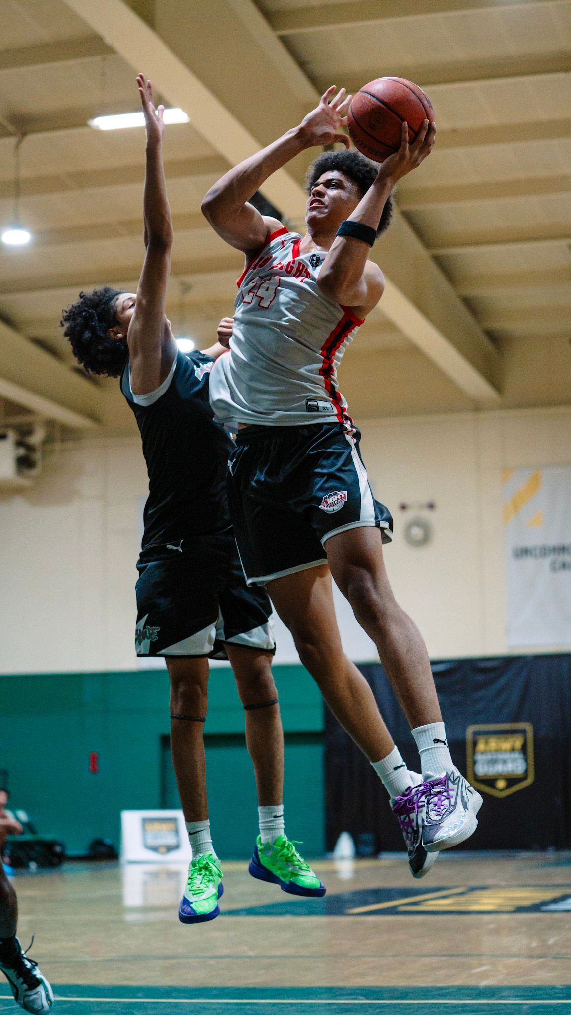Basketball player in mid-air, shooting, defended by an opponent. Indoor court setting.