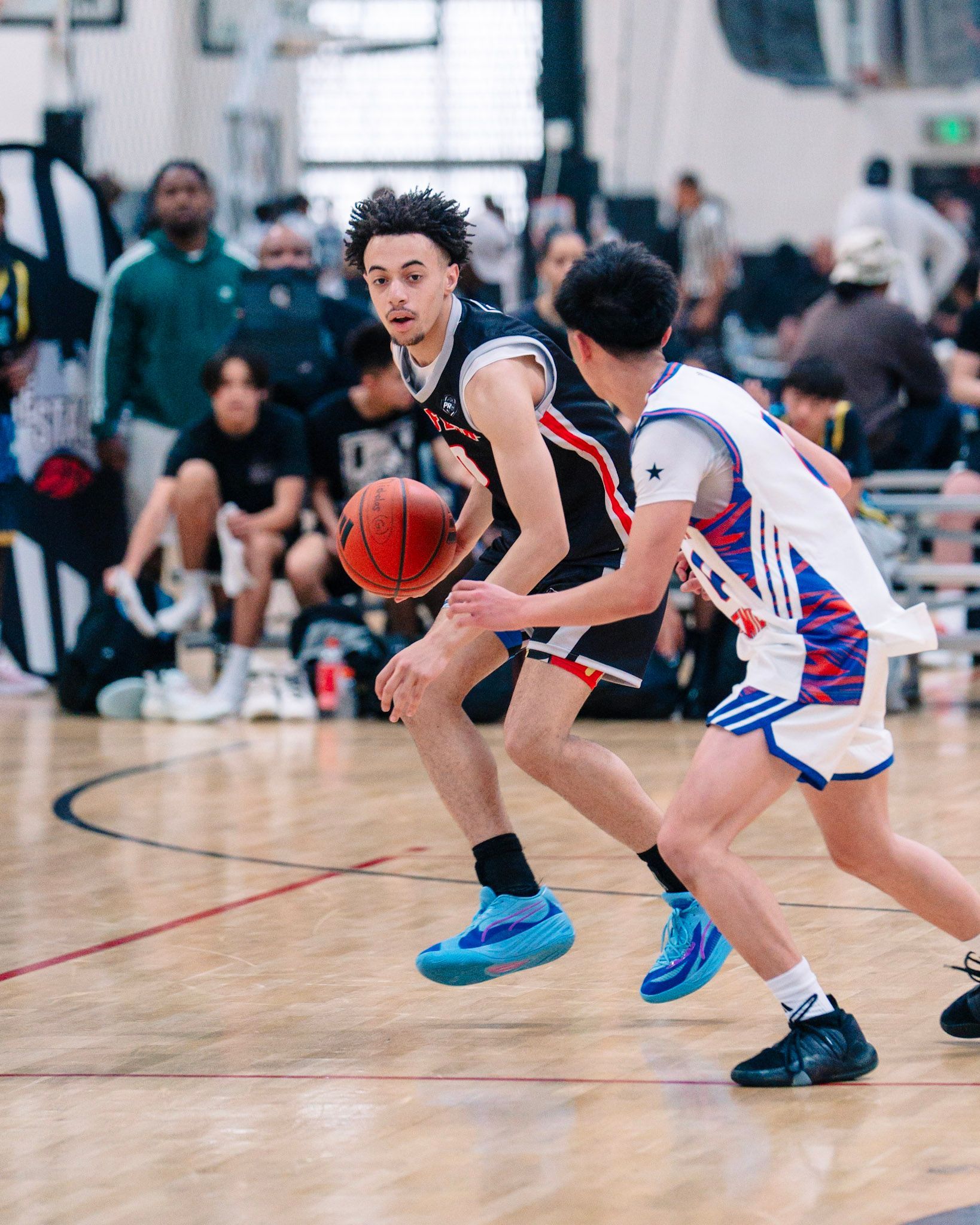 Basketball players face off on court; one dribbles, guarded closely.