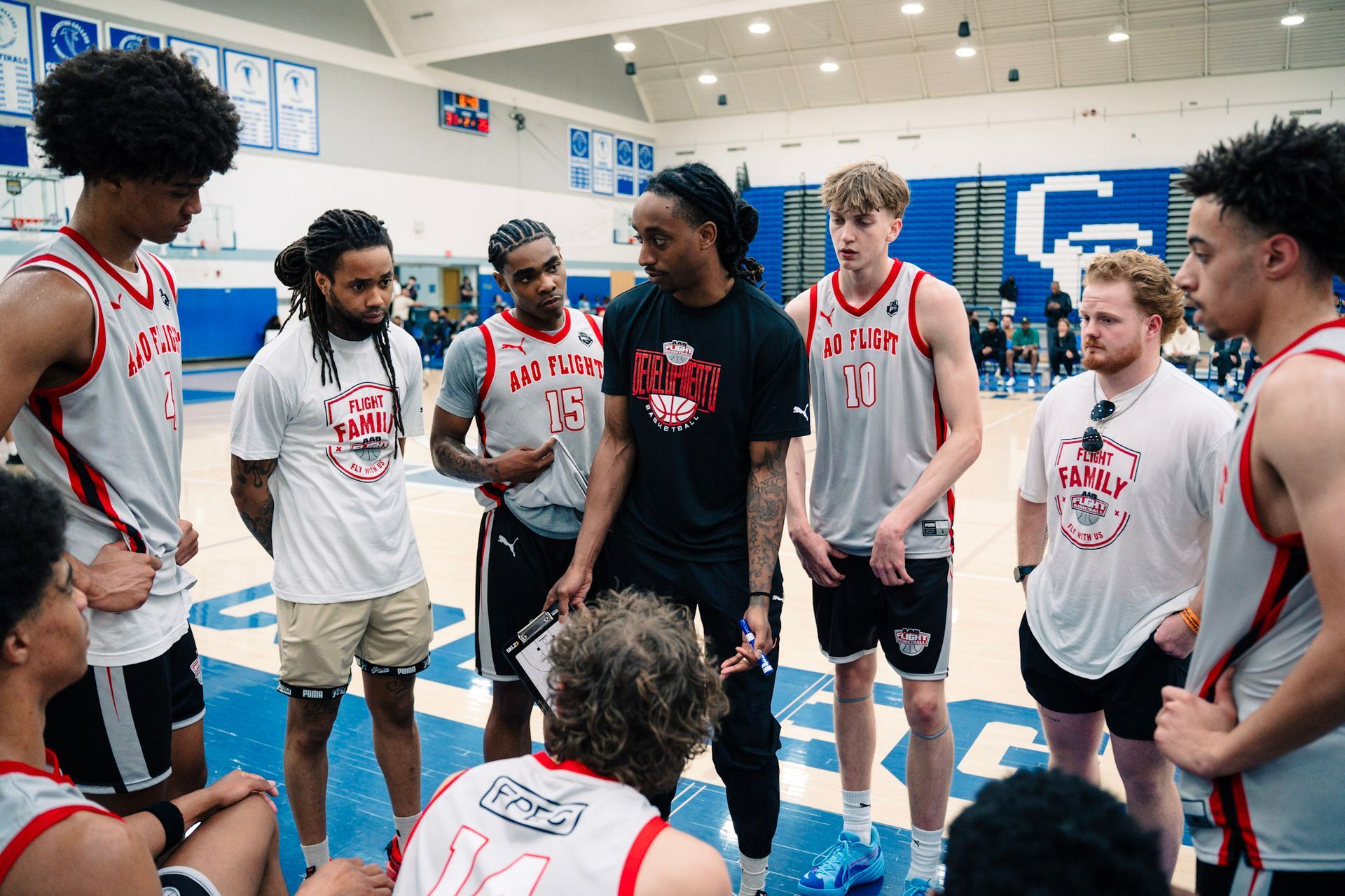 Basketball coach talking to team on a court. Players in red & white jerseys, listening intently. Blue court background.
