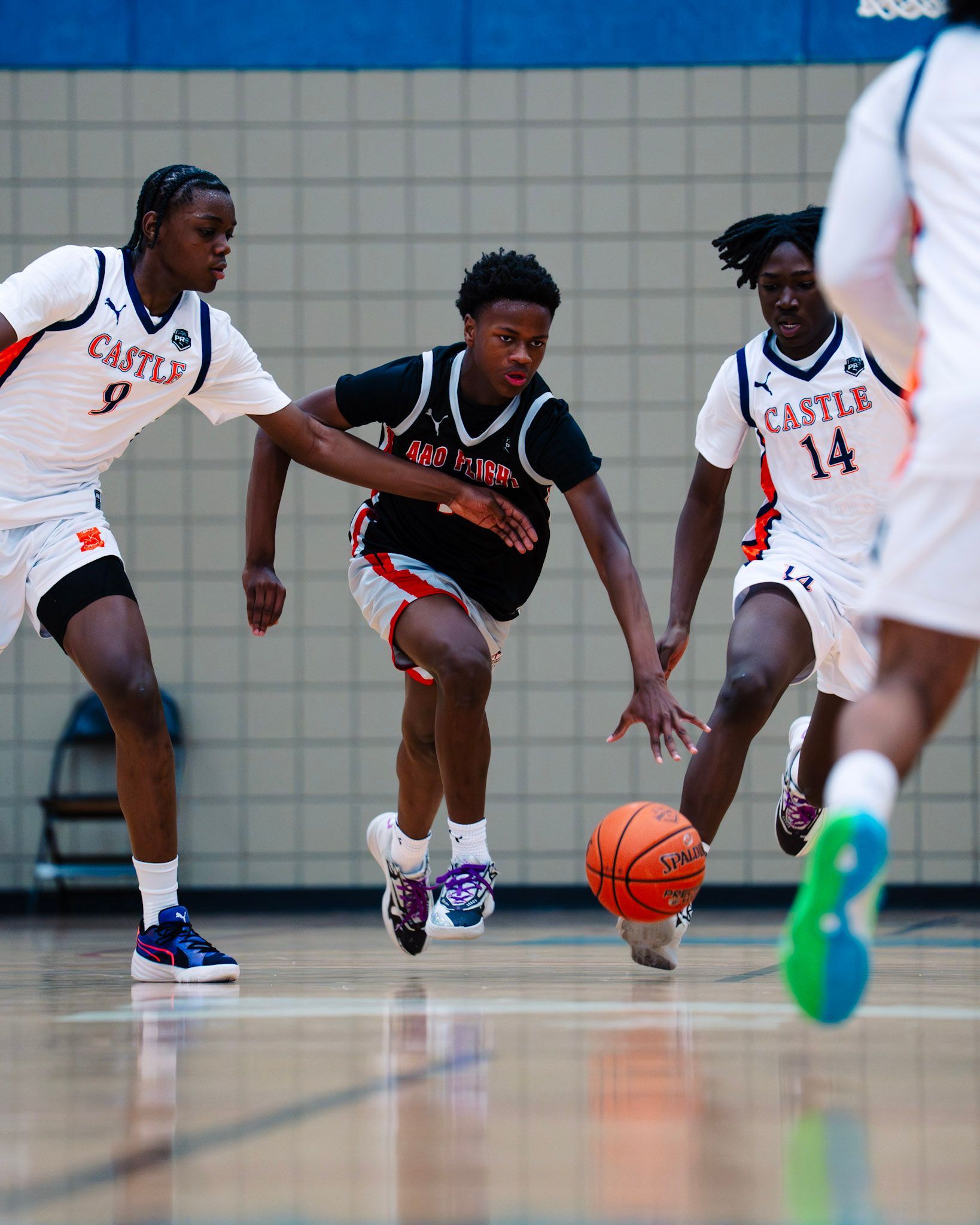 Basketball game: Player dribbling ball, pursued by two defenders in white and orange jerseys on court.