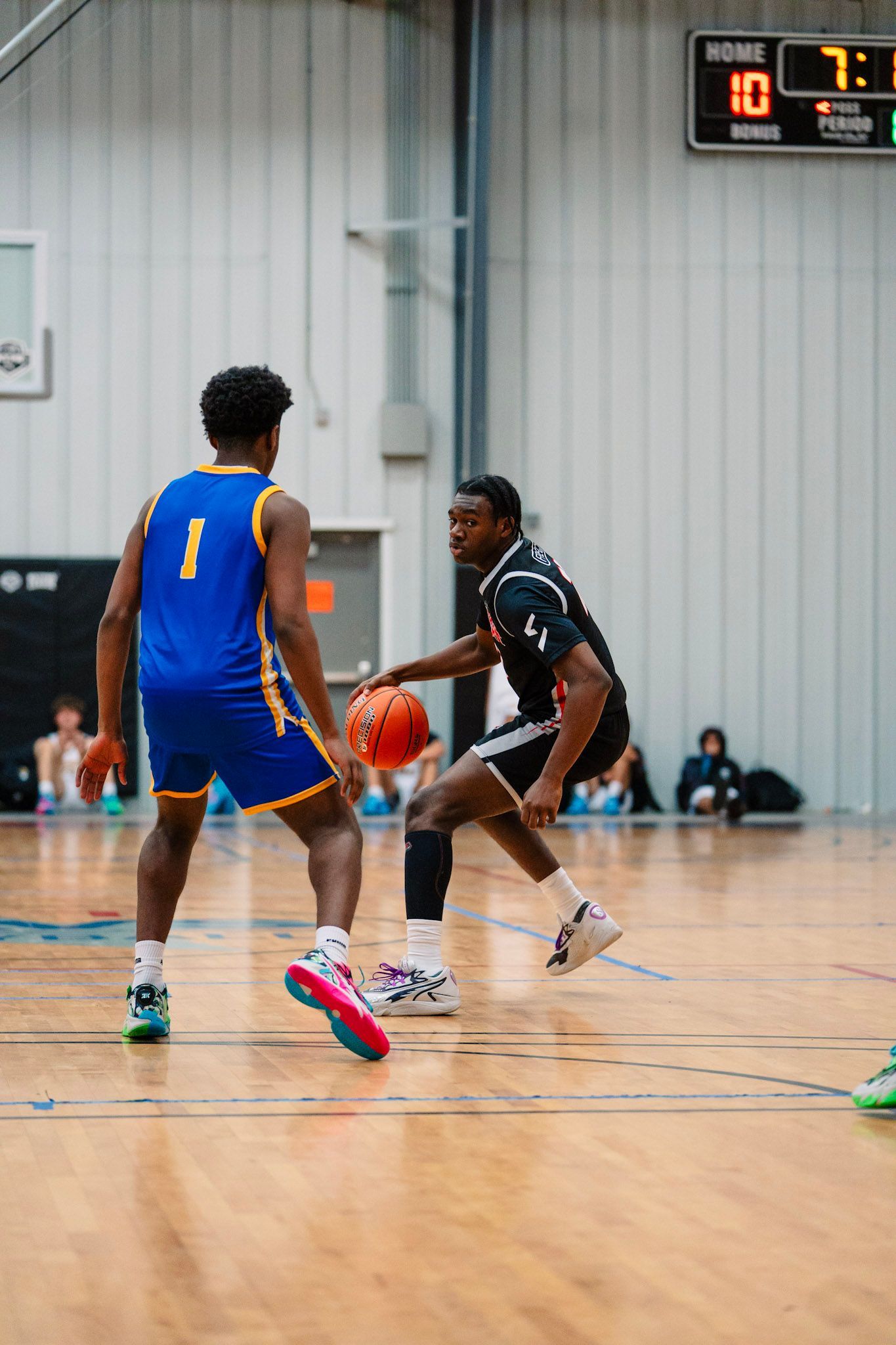 Basketball game: Player in black dribbles past a player in blue; scoreboard visible.