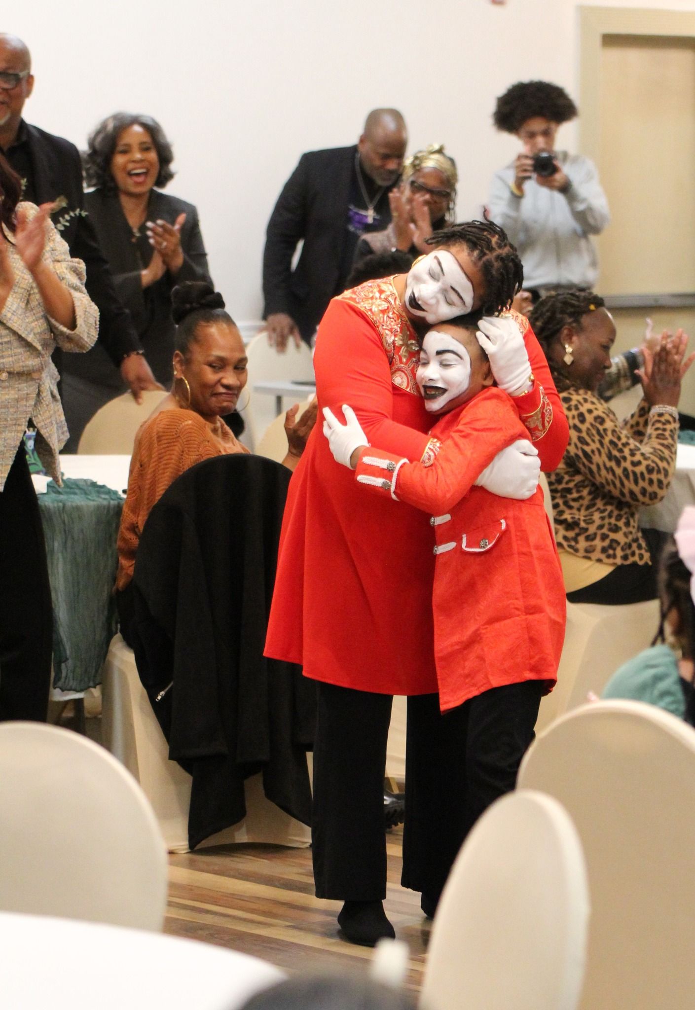 A woman in a red coat is hugging a child with white face paint