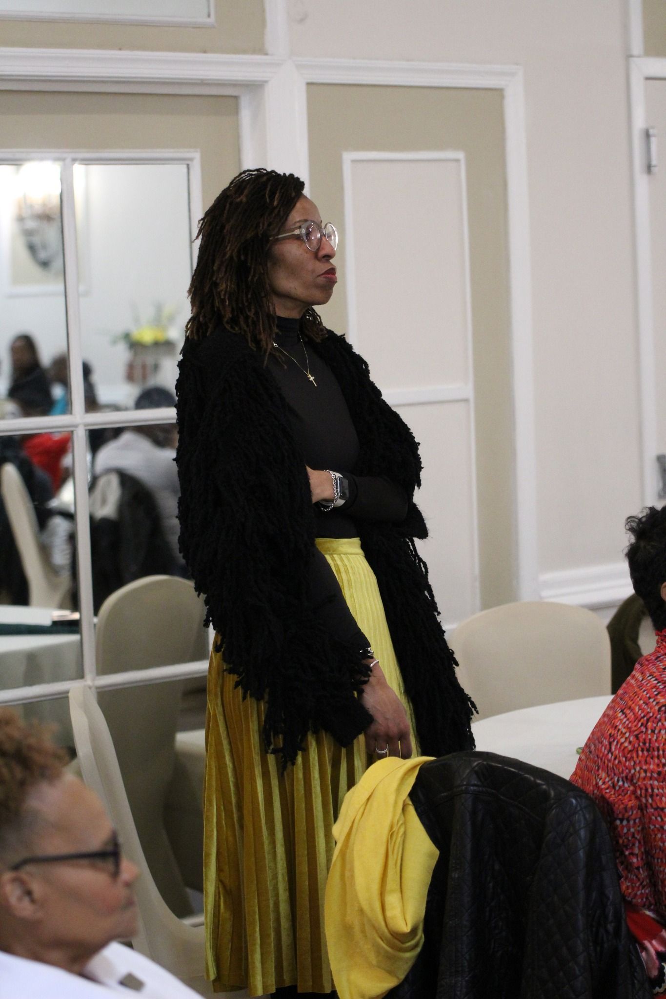 A woman in a black coat and yellow skirt is standing in front of a group of people.