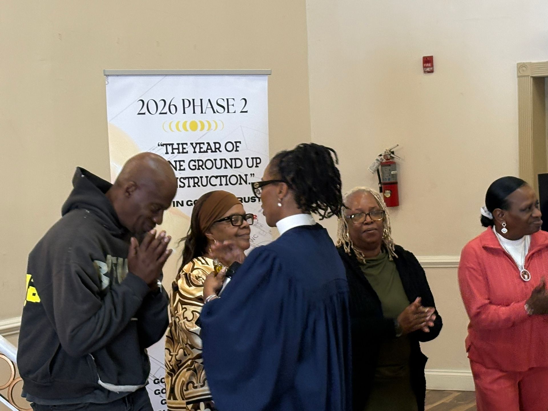 People gathered indoors for a meeting, standing near a sign reading “2026 Phase 2.”