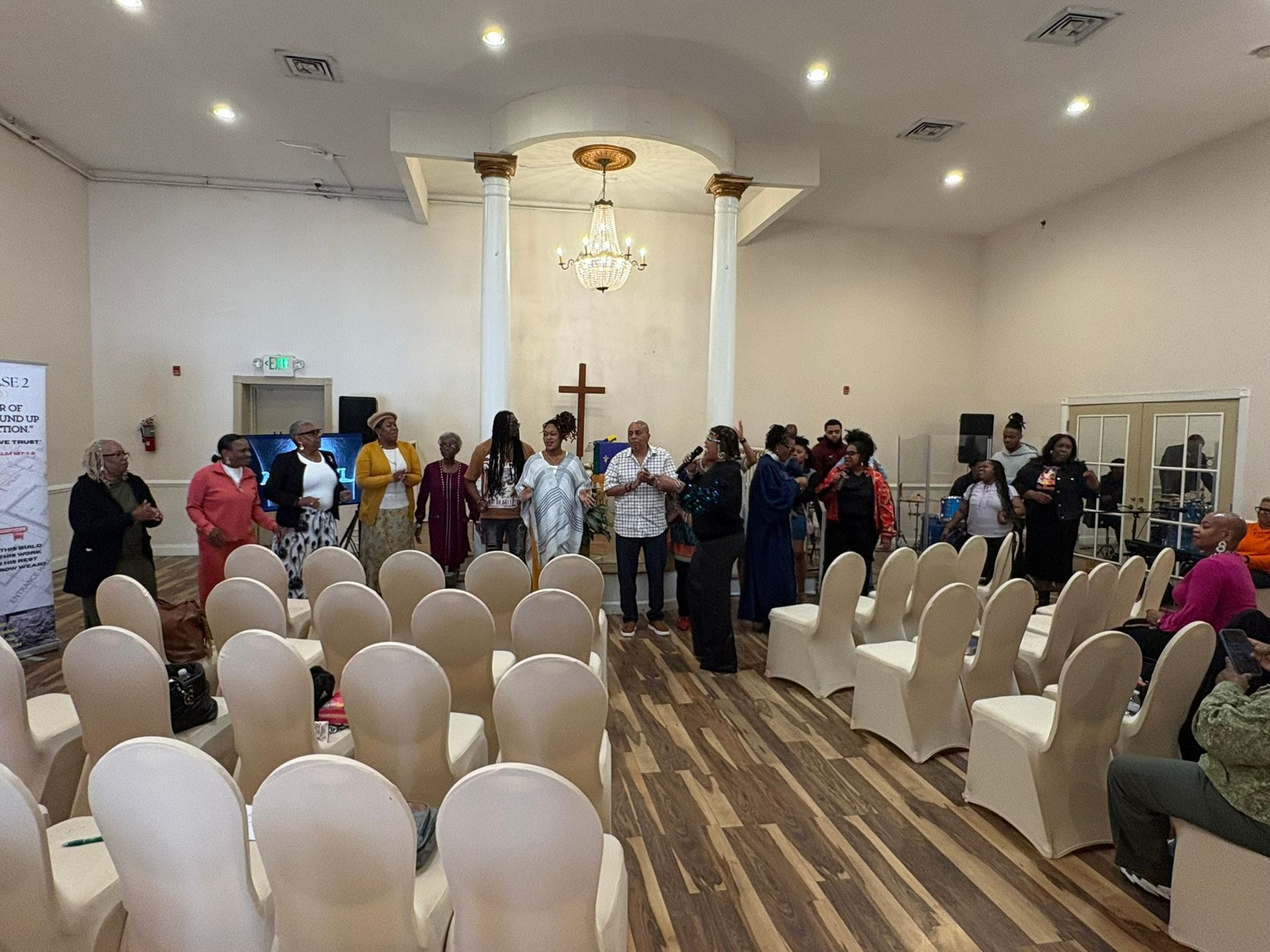 People standing at the front of a chapel with rows of white chairs and a cross on the altar